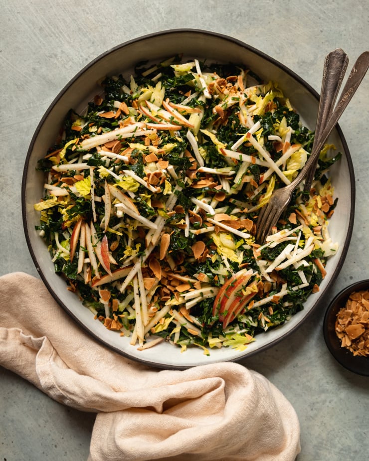 An overhead shot of an apple kale salad with toasted almonds, photographed in a white ceramic dish that is rimmed with matte black. A pale yellow linen is seen to the side.