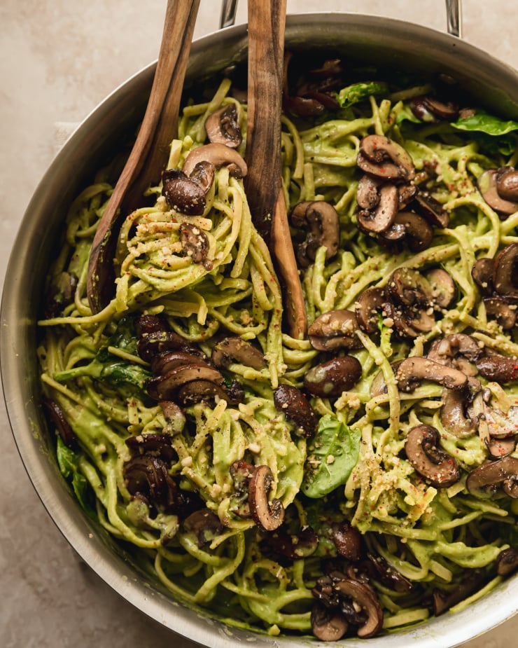 pinach and mushroom pasta in a stainless steel skillet. The pasta is creamy and pale green and topped with brown, sautéed mushrooms. A pair of wooden serving tongs are visible.