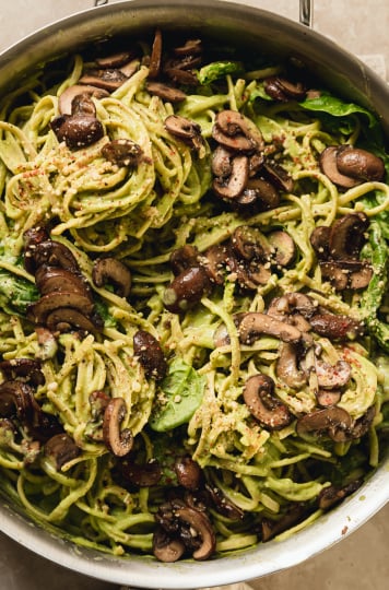 spinach and mushroom pasta in a stainless steel skillet. The pasta is creamy and pale green and topped with brown, sautéed mushrooms.