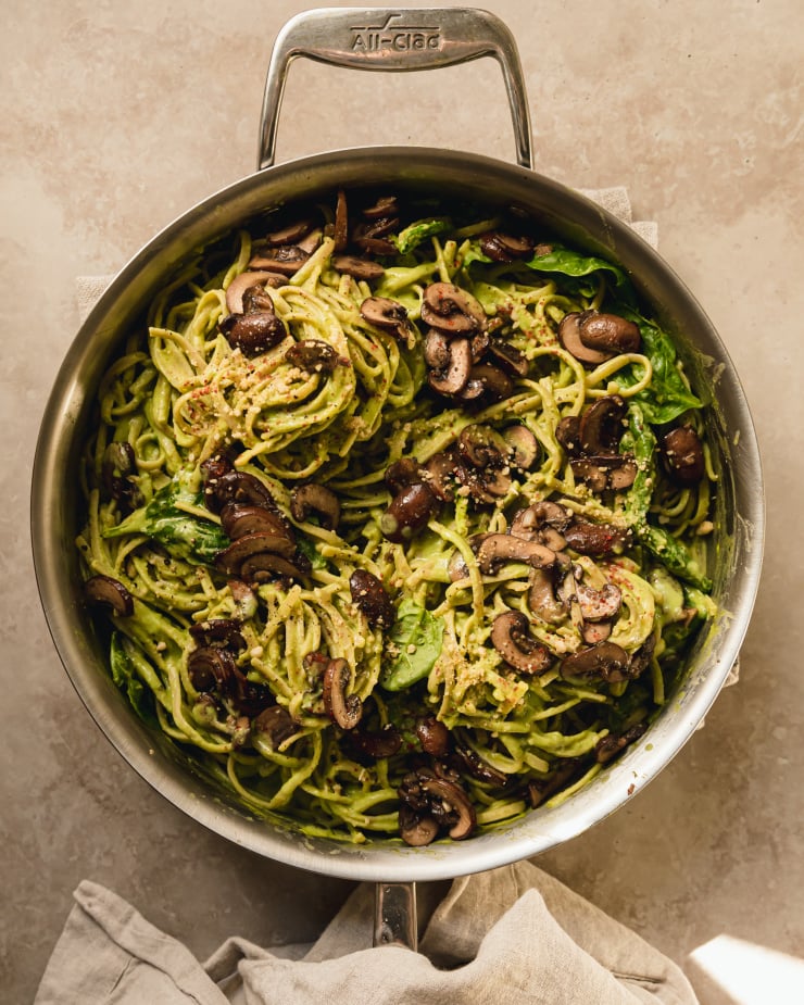 An overhead shot of a creamy vegan spinach and mushroom pasta in a stainless steel skillet. The pasta is creamy and pale green and topped with brown, sautéed mushrooms.