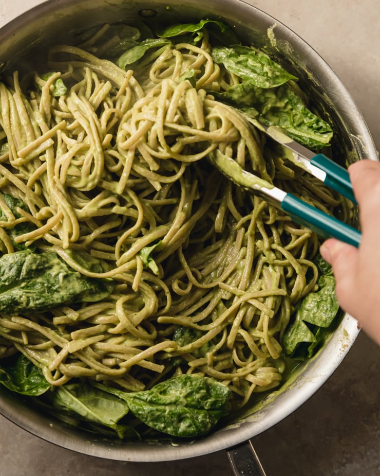 An overhand shot of a hand using tongs to toss together some cooked linguine, baby spinach, and a creamy sauce.