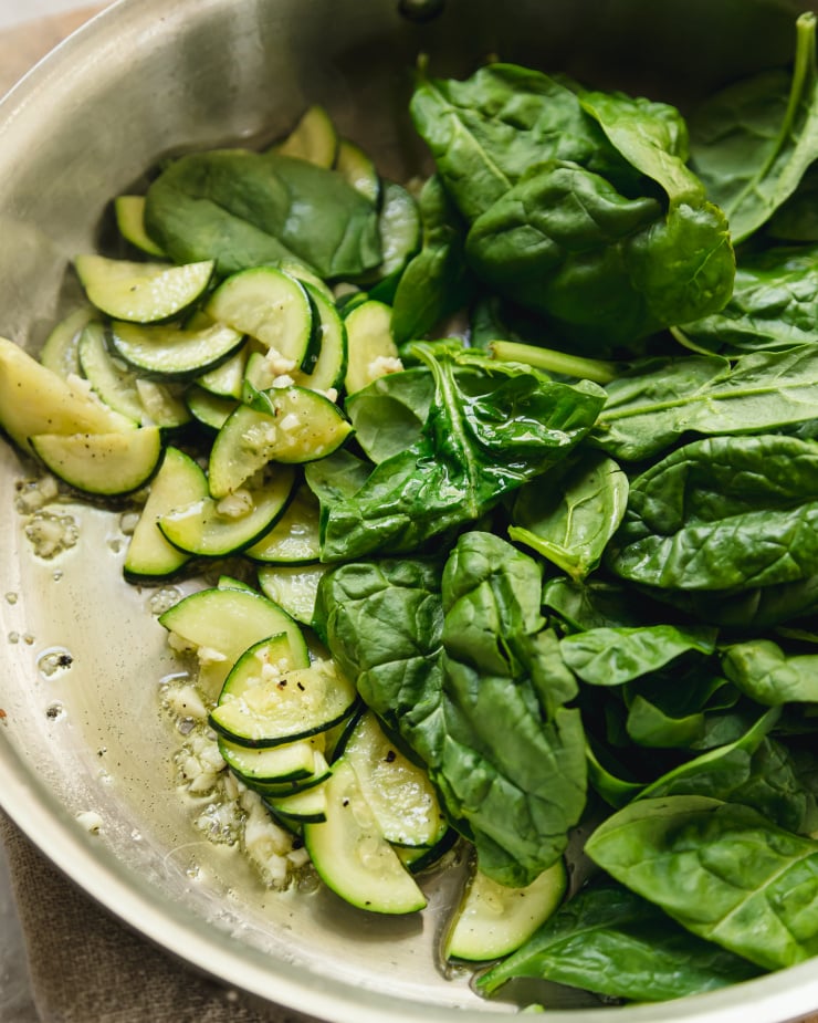 An up close, 3/4 angle shot of zucchini slices and spinach sautéing in a stainless steel pan.