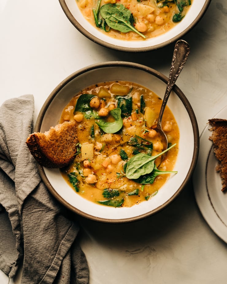 An overhead shot of a creamy and thick lemony chickpea soup with spinach and potatoes in a white ceramic bowl edges with grey. A piece of whole grain bread is sticking out of the soup.