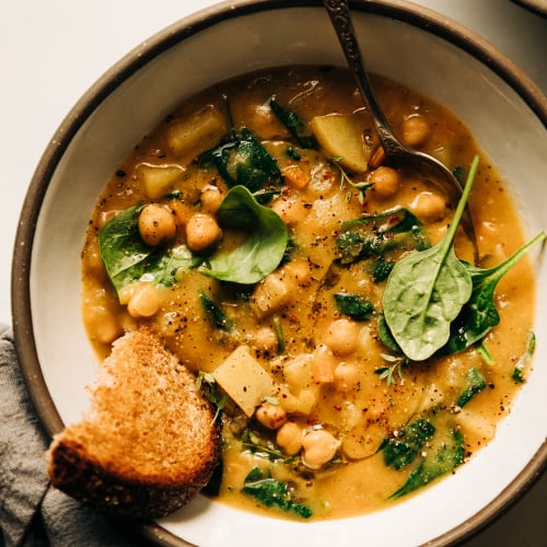 An overhead shot of a creamy and thick lemony chickpea soup with spinach and potatoes in a white ceramic bowl edges with grey. A piece of whole grain bread is sticking out of the soup.