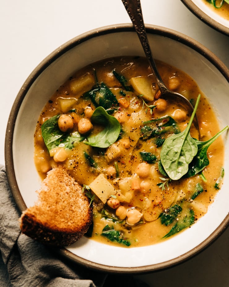 An overhead shot of a creamy and thick lemony chickpea soup with spinach and potatoes in a white ceramic bowl edges with grey. A piece of whole grain bread is sticking out of the soup.