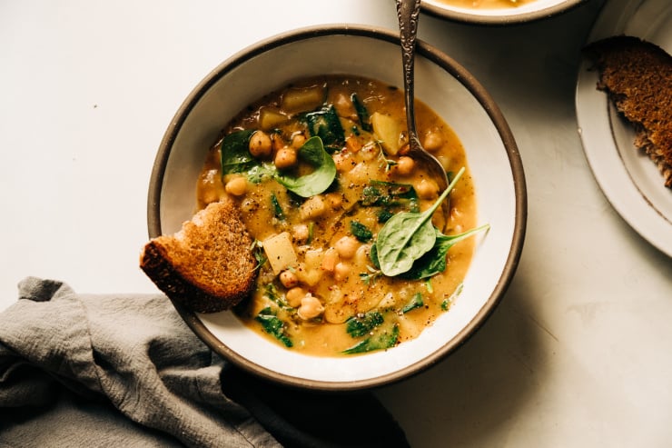 An overhead shot of a creamy and thick lemony chickpea soup with spinach and potatoes in a white ceramic bowl edges with grey. A piece of whole grain bread is sticking out of the soup.