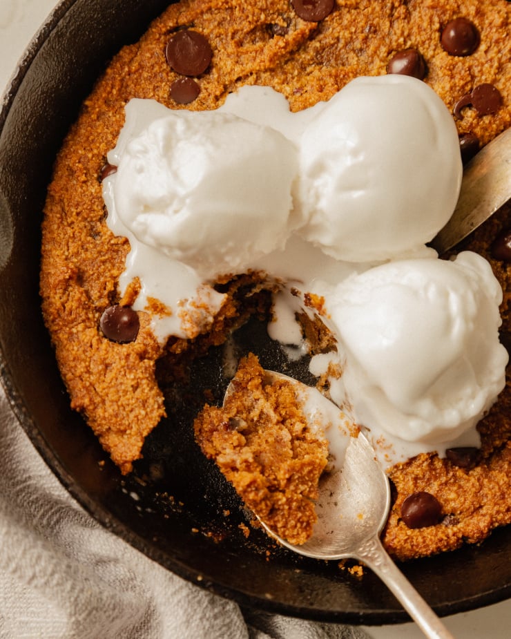 An up close, overhead shot of a slightly orange pumpkin chocolate chip skillet cookie topped with 3 scoops of vanilla ice cream. The ice cream is melt-y and several bites have been taken out of the cookie.