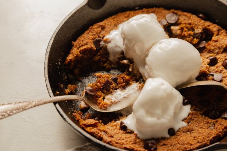 A 3/4 angle shot of a slightly orange pumpkin chocolate chip skillet cookie topped with 3 scoops of vanilla ice cream. The ice cream is melt-y and several bites have been taken out of the cookie.
