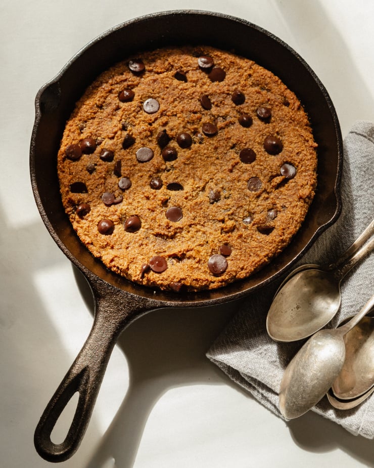 An overhead shot of a large cookie that has been baked in a skillet. There is a grey napkin with a few spoons on top nearby.