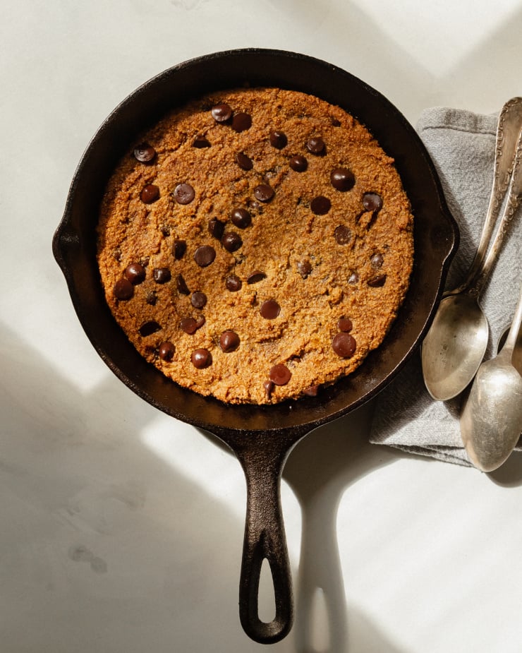 An overhead shot of a large cookie that has been baked in a skillet. There is a grey napkin with a few spoons on top nearby.