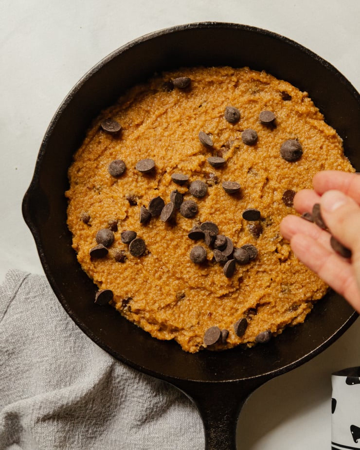 A hand is seen sprinkling chocolate chips on top of a large portion of cookie dough that is contained in a cast iron skillet.