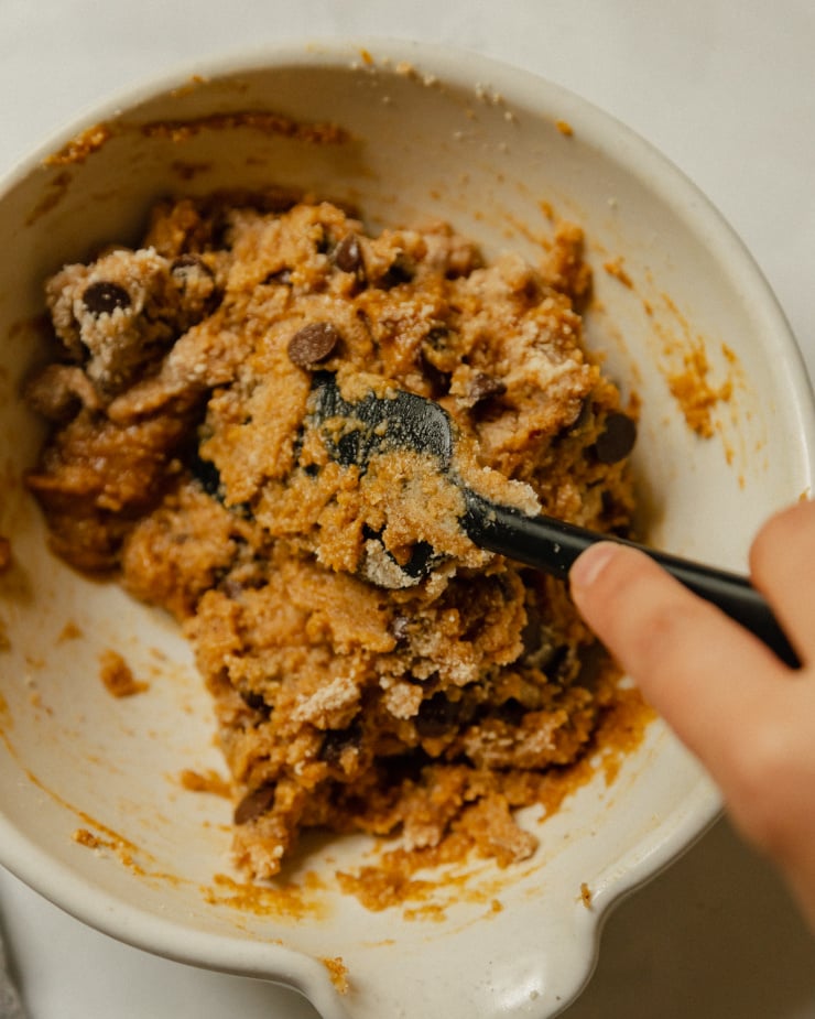 A hand is folding together some cookie dough with a spatula in a cream coloured mixing bowl.