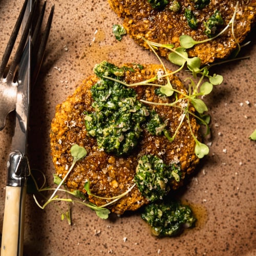 An overhead shot of a butternut squash patty on a speckled brown plate topped with deep green salsa verde and arugula sprouts.