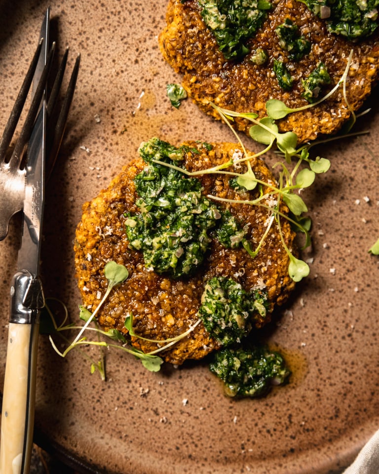 An overhead shot of a butternut squash patty on a speckled brown plate topped with deep green salsa verde and arugula sprouts.