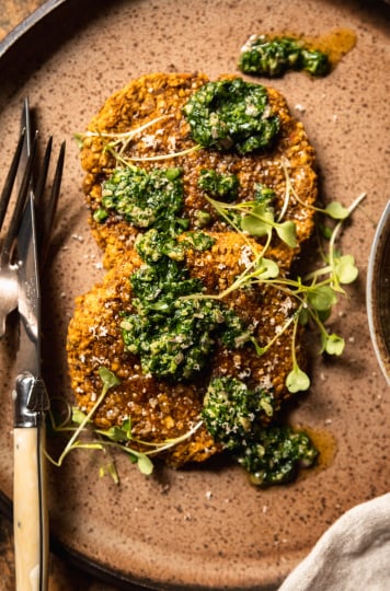 An overhead shot of two vegan squash patties on a speckled brown plate topped with deep green salsa verde and arugula sprouts.