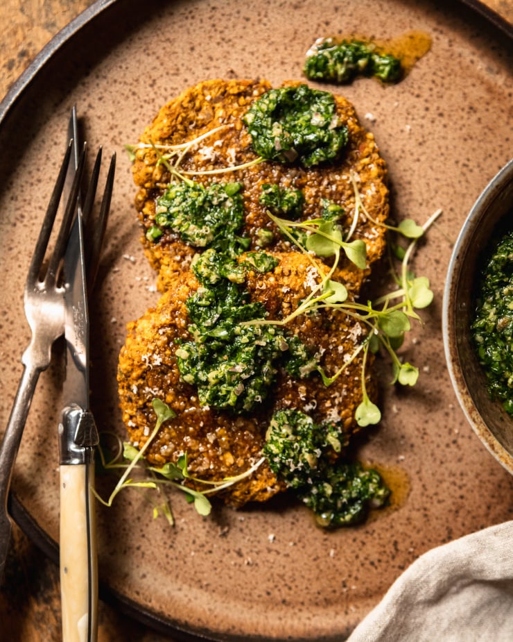 An overhead shot of two vegan squash patties on a speckled brown plate topped with deep green salsa verde and arugula sprouts.