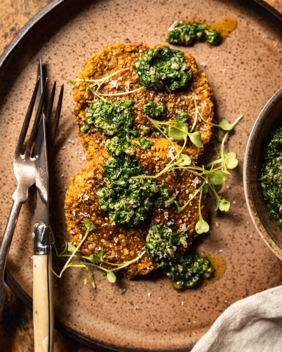 An overhead shot of two vegan squash patties on a speckled brown plate topped with deep green salsa verde and arugula sprouts.