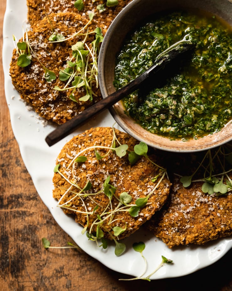 An overhead shot of vegan squash patties on a white platter with deep green mixed herb salsa verde in a bowl to the side. The patties are topped with arugula sprouts and fine shavings of pecans.