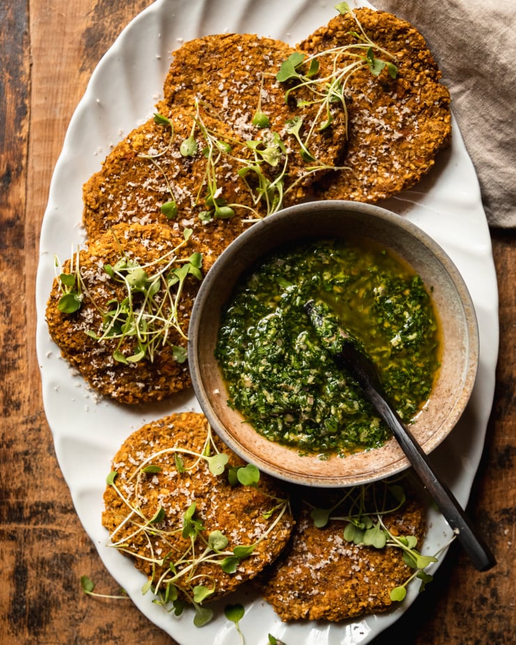An overhead shot of vegan squash patties on a white platter with deep green mixed herb salsa verde in a bowl to the side. The patties are topped with arugula sprouts and fine shavings of pecans.