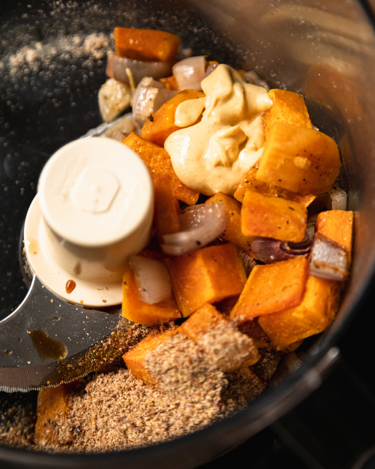 An overhead shot of roasted squash, shallots, and flax meal in the bowl of a food processor.