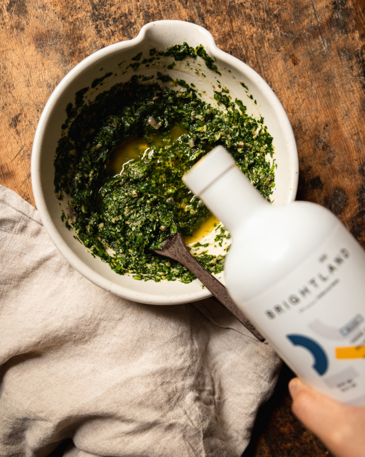 Image shows a hand pouring olive oil into a small bowl of mixed salsa verde on a wood background.