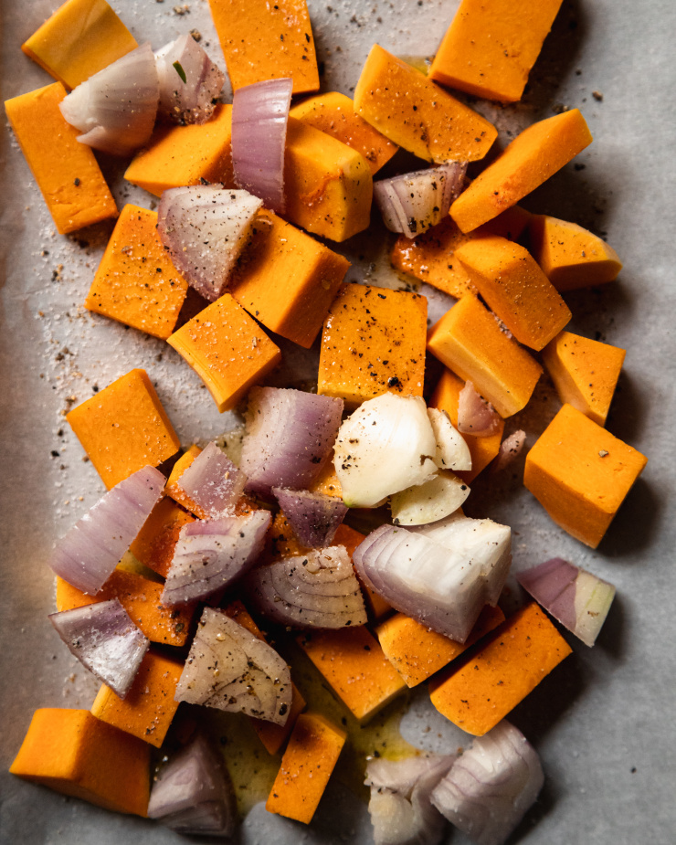 An up close, overhead image of squash cubes, diced shallots and a garlic clove on a parchment lined baking sheet.