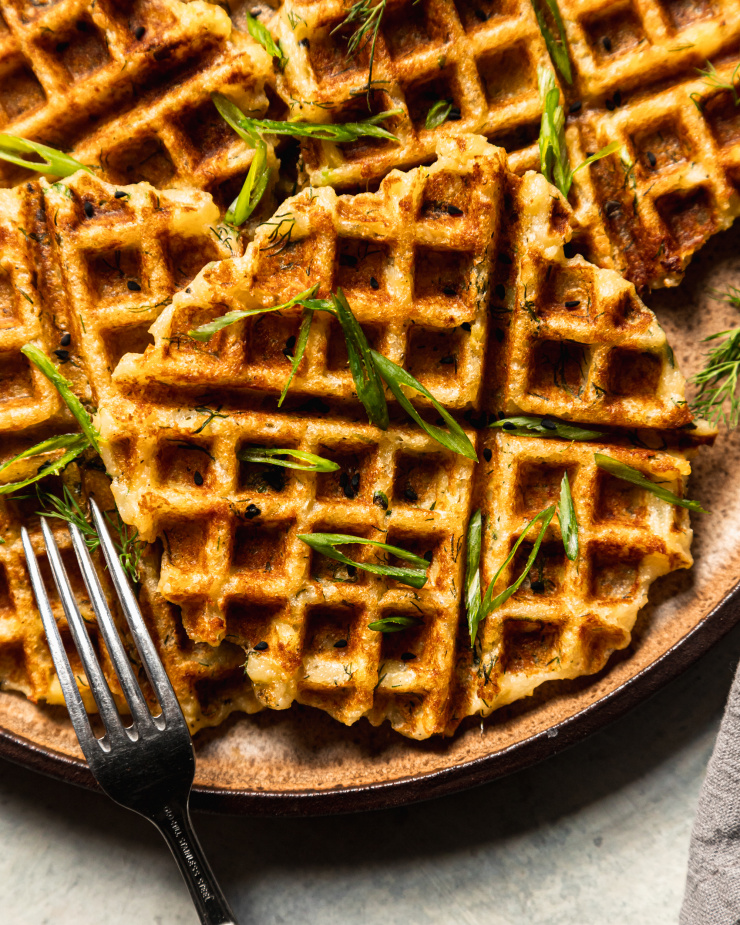 An overhead shot of crisp and golden waffles on a brown/tan flecked plate. The waffles are topped with sliced green onions, nigella seeds, and chopped dill.