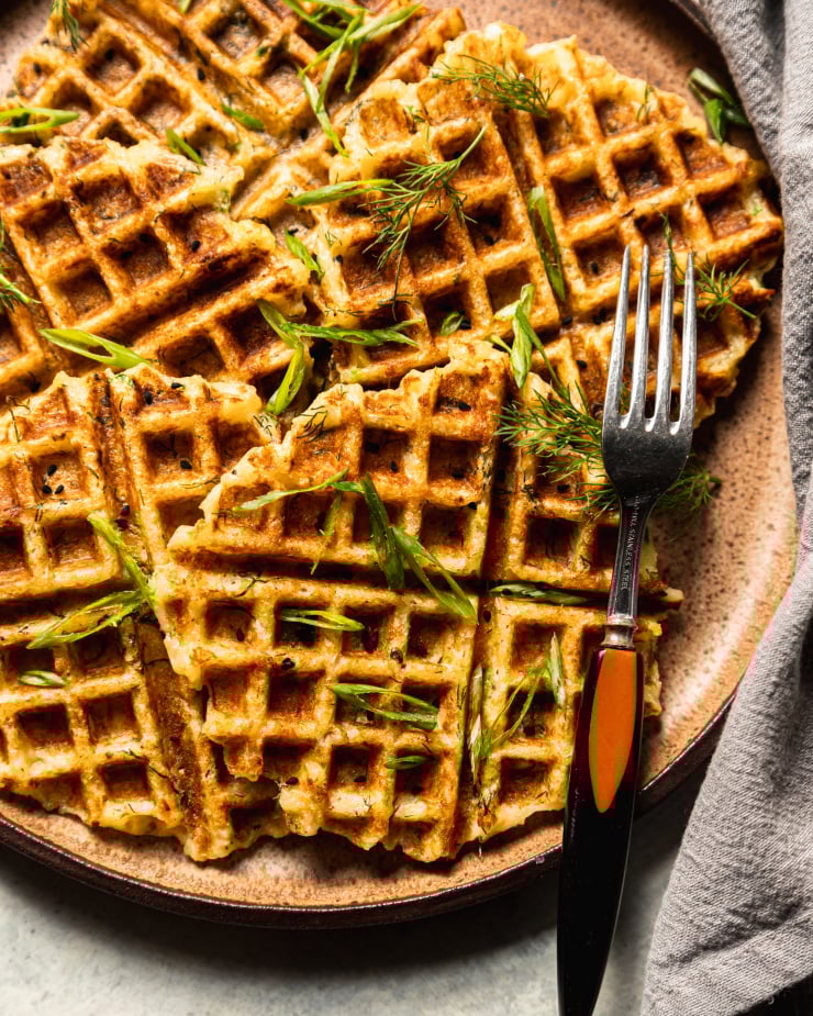 An overhead shot of crisp and golden waffles on a brown/tan flecked plate. The waffles are topped with sliced green onions, nigella seeds, and chopped dill.