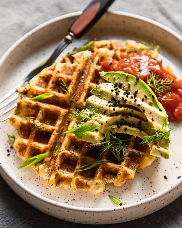 An overhead shot of two crispy golden waffles on a white plate topped with salsa and sliced avocado. A fork is also on the plate to the side.