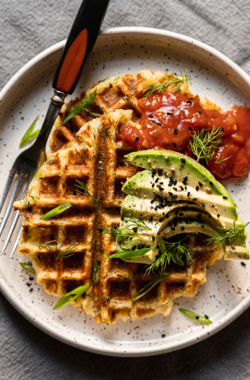 An overhead shot of two crispy golden waffles on a white plate topped with salsa and sliced avocado. A fork is also on the plate to the side.