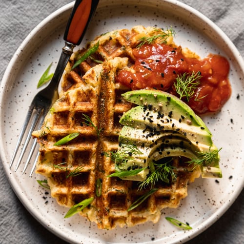 An overhead shot of two crispy golden waffles on a white plate topped with salsa and sliced avocado. A fork is also on the plate to the side.