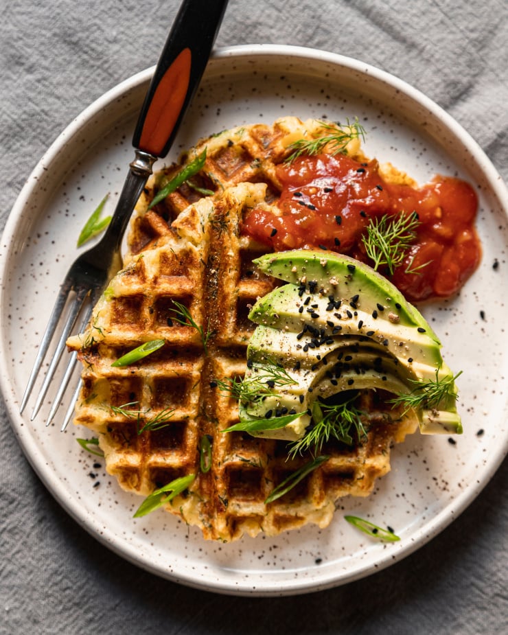 An overhead shot of two crispy golden waffles on a white plate topped with salsa and sliced avocado. A fork is also on the plate to the side.