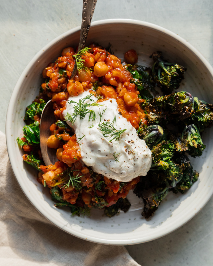 An overhead shot of a farro, chickpea and tomato stew in a grey-ish white bowl. The stew is served with a dollop of bright white yogurt sauce on top, chopped dill, and roasted kalettes to the side.
