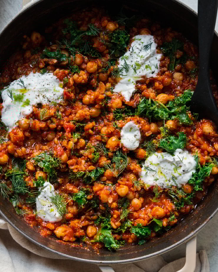 An up close, overhead shot of a farro chickpea skillet with crushed tomatoes, chopped kale, and dollops of a vegan yogurt sauce on top. There is a black, wooden spoon sticking out of the skillet.