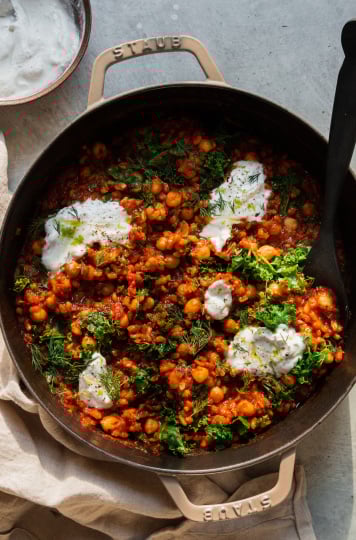 An overhead shot of a farro chickpea skillet with crushed tomatoes, chopped kale, and dollops of a vegan yogurt sauce on top. There is a black, wooden spoon sticking out of the skillet.