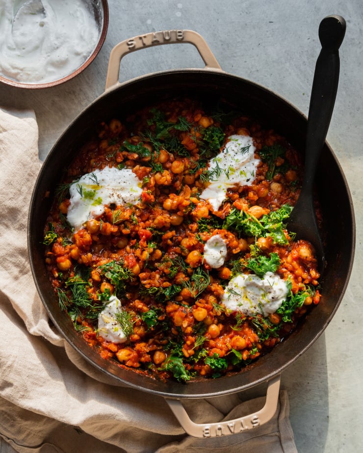 An overhead shot of a farro chickpea skillet with crushed tomatoes, chopped kale, and dollops of a vegan yogurt sauce on top. There is a black, wooden spoon sticking out of the skillet.