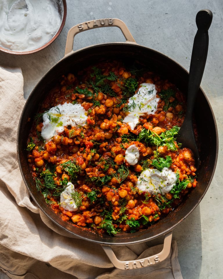 An overhead shot of a farro chickpea skillet with crushed tomatoes, chopped kale, and dollops of a vegan yogurt sauce on top. There is a black, wooden spoon sticking out of the skillet.