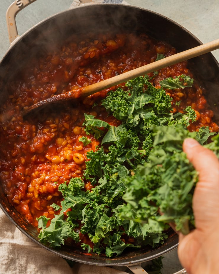 An overhead shot of a hand adding chopped kale to a farro chickpea skillet with tomatoes. The mixture is steaming.