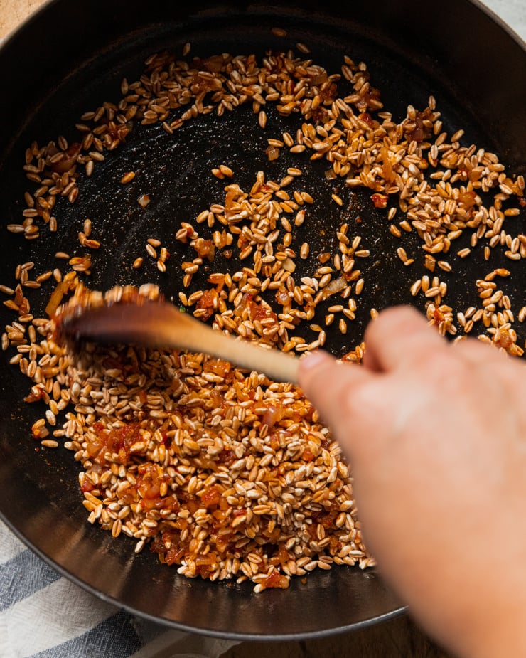 An overhead shot of a hand using a wooden spoon to stir up grains of farro in spices within a pan.