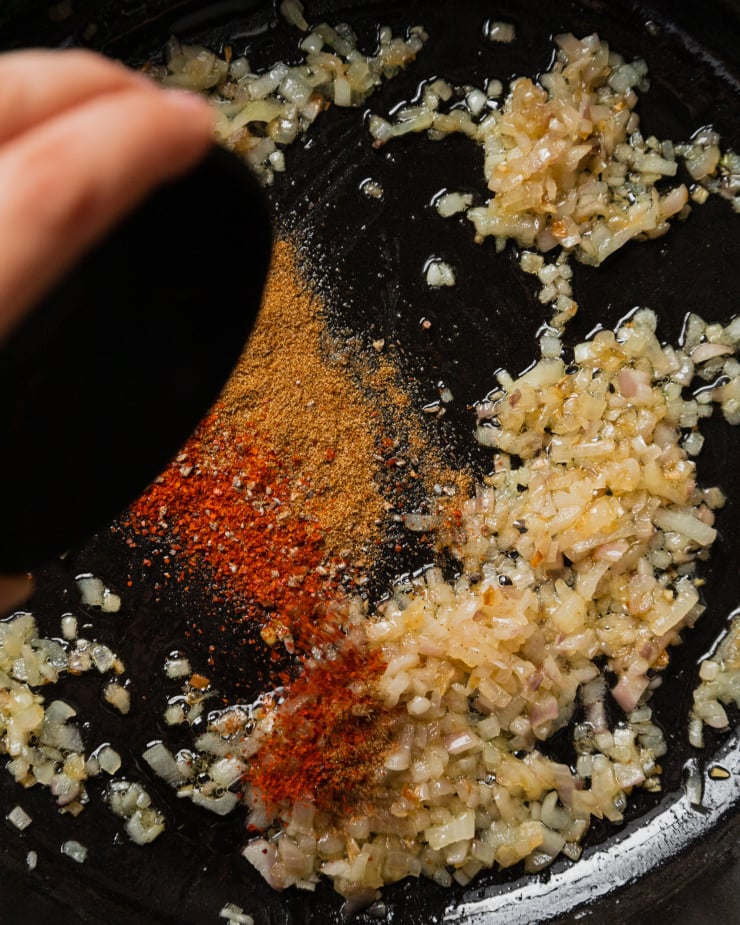 An up close, overhead shot of a hand pouring a bowl of spices into a pan with sautéed shallots.