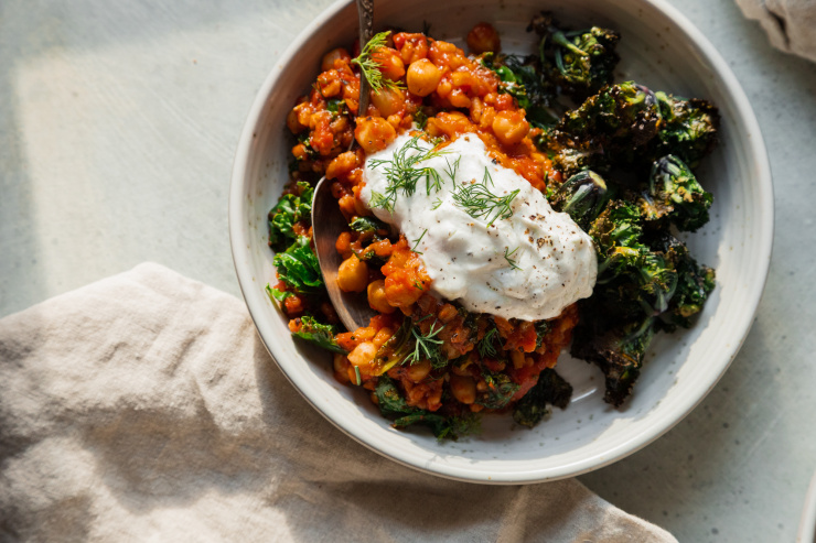 An overhead shot of a farro, chickpea and tomato stew in a grey-ish white bowl. The stew is served with a dollop of bright white yogurt sauce on top, chopped dill, and roasted kalettes to the side.