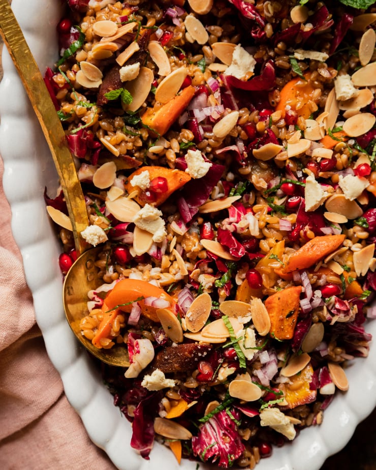 An overhead shot of a vegan holiday farro salad topped with fresh mint, roasted carrots, chopped radicchio, pomegranate arils, slices of persimmon, and toasted sliced almonds. The salad is in a white, scalloped edge oval dish with a brass serving ladle.