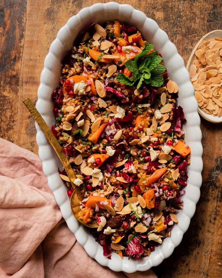 An overhead shot of a vegan holiday farro salad topped with fresh mint, roasted carrots, chopped radicchio, pomegranate arils, slices of persimmon, and toasted sliced almonds. The salad is in a white, scalloped edge oval dish with a brass serving ladle.