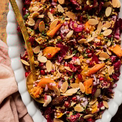 An overhead shot of a vegan holiday farro salad topped with fresh mint, roasted carrots, chopped radicchio, pomegranate arils, slices of persimmon, and toasted sliced almonds. The salad is in a white, scalloped edge oval dish with a brass serving ladle.