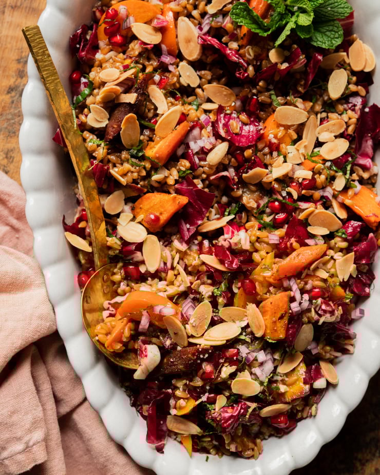 An overhead shot of a vegan holiday farro salad topped with fresh mint, roasted carrots, chopped radicchio, pomegranate arils, slices of persimmon, and toasted sliced almonds. The salad is in a white, scalloped edge oval dish with a brass serving ladle.