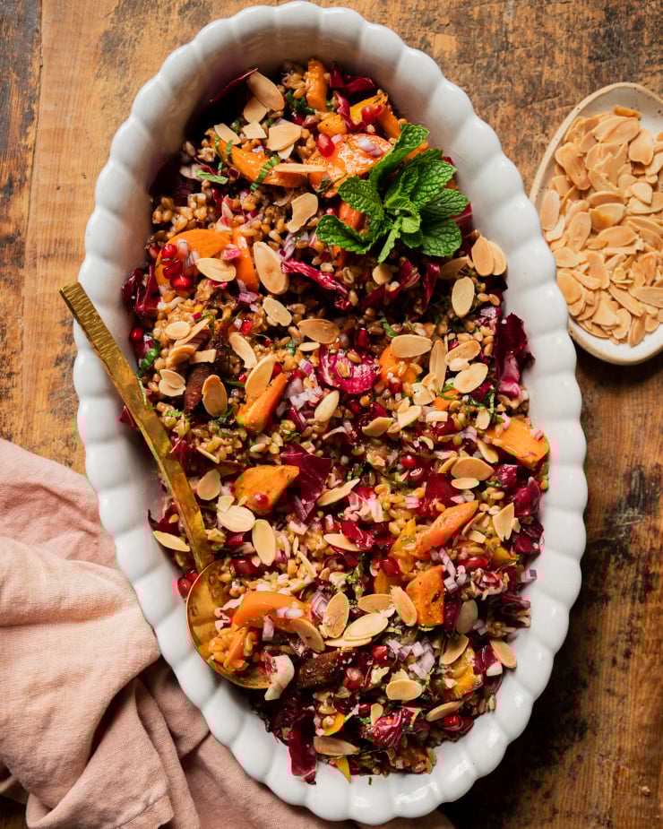 An overhead shot of a vegan holiday farro salad topped with fresh mint, roasted carrots, chopped radicchio, pomegranate arils, slices of persimmon, and toasted sliced almonds. The salad is in a white, scalloped edge oval dish with a brass serving ladle.