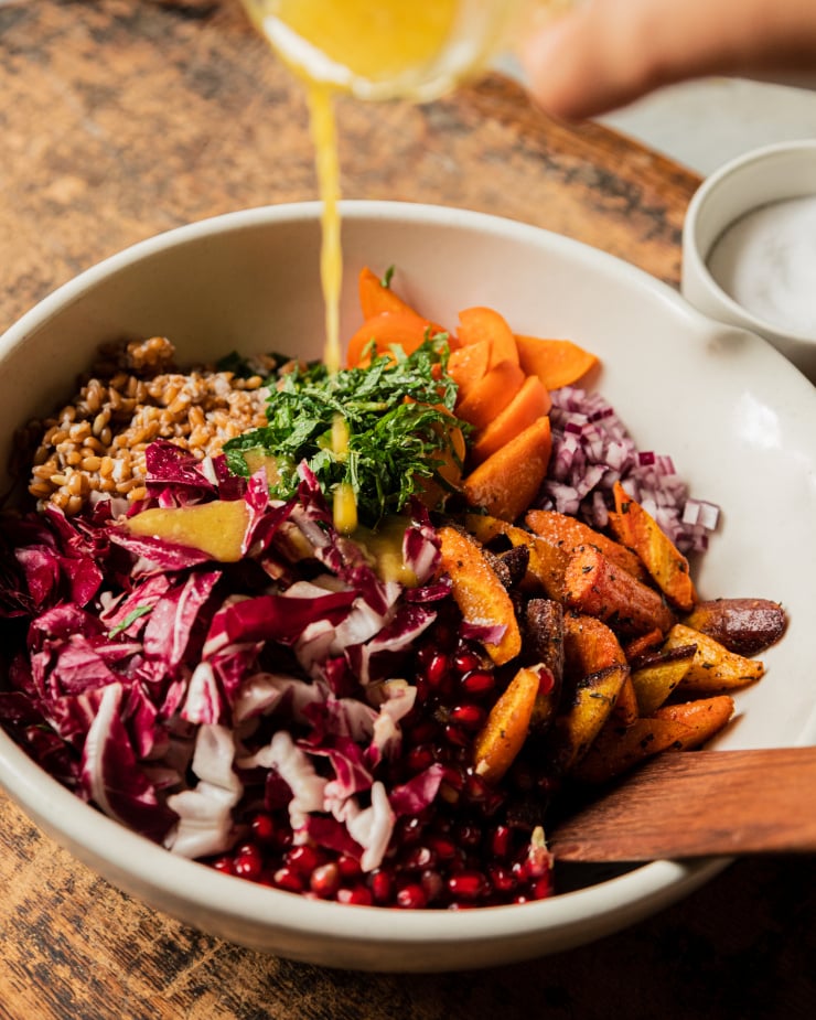 A 3/4 angle shot of dressing being poured into a white mixing bowl filled with components of a farro salad, pre-mixing.