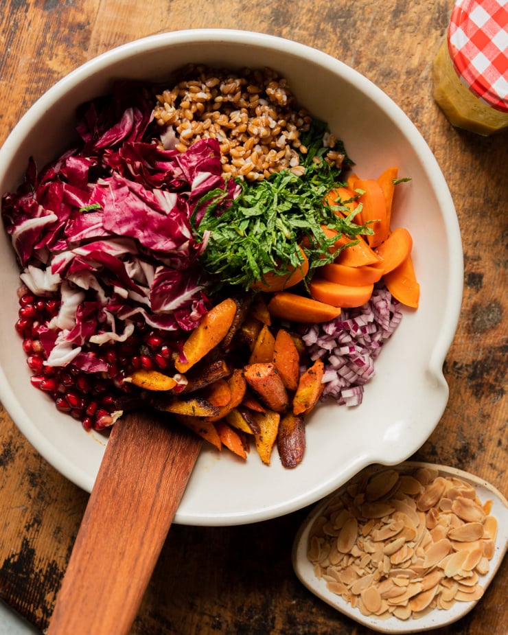An overhead shot of all prepped components of a holiday farro salad in an off white mixing bowl with a wooden spatula sticking out.