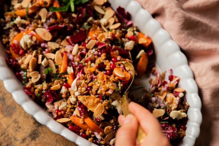 A 3/4 angle shot of a hand spooning out a portion of farro salad that's topped with roasted carrots, pomegranate seeds, sliced almonds, and chopped radicchio.