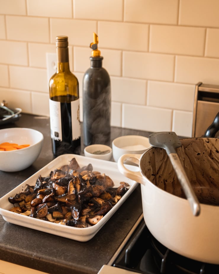 A 3/4 angle image shows sautรฉed mushrooms in a white dish on a kitchen counter next to a gas stove. A bottle of red wine is in the background.